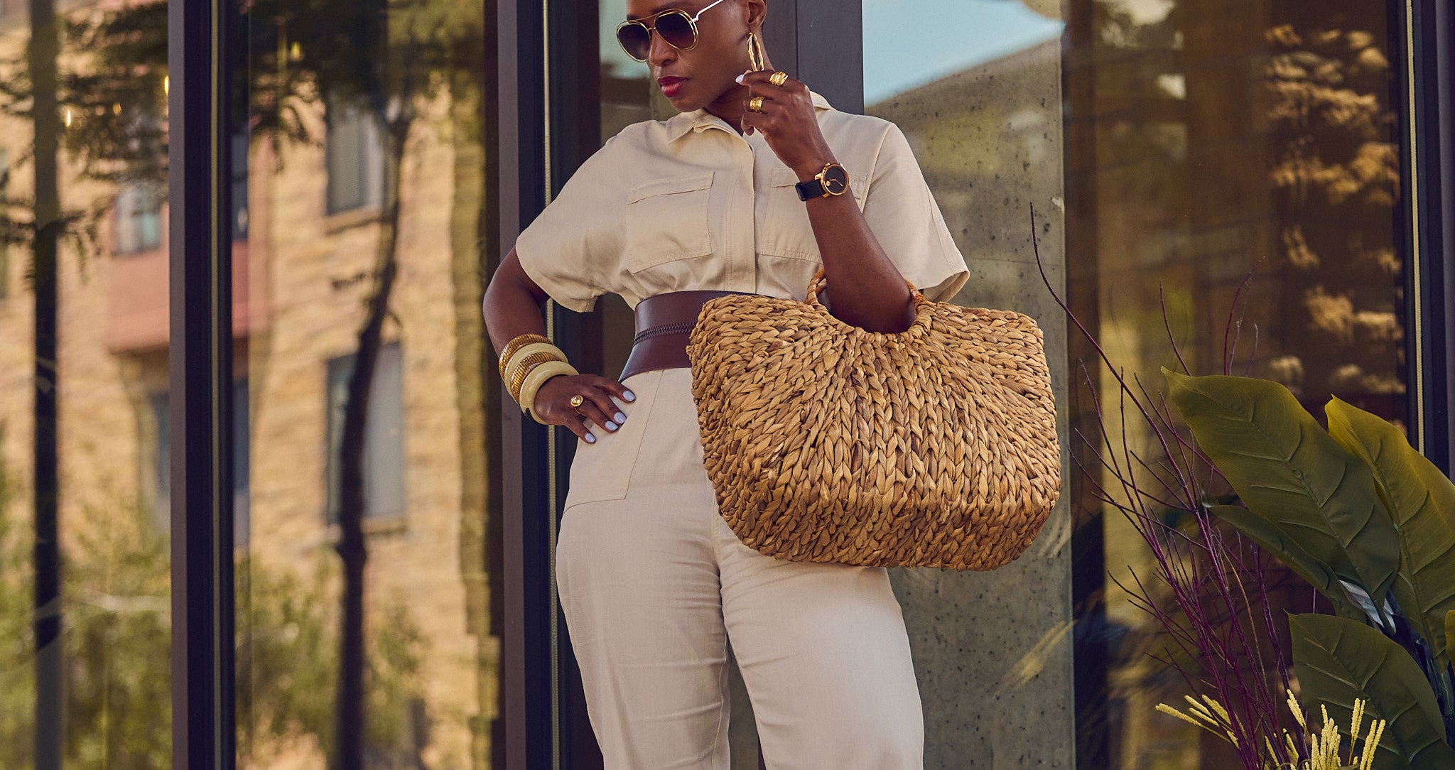 Influencer Farotelle standing against a wall and wearing a light beige jumpsuit with a wide brown waist belt, sunglasses, chunky bangle bracelets, and holding a large straw bag.