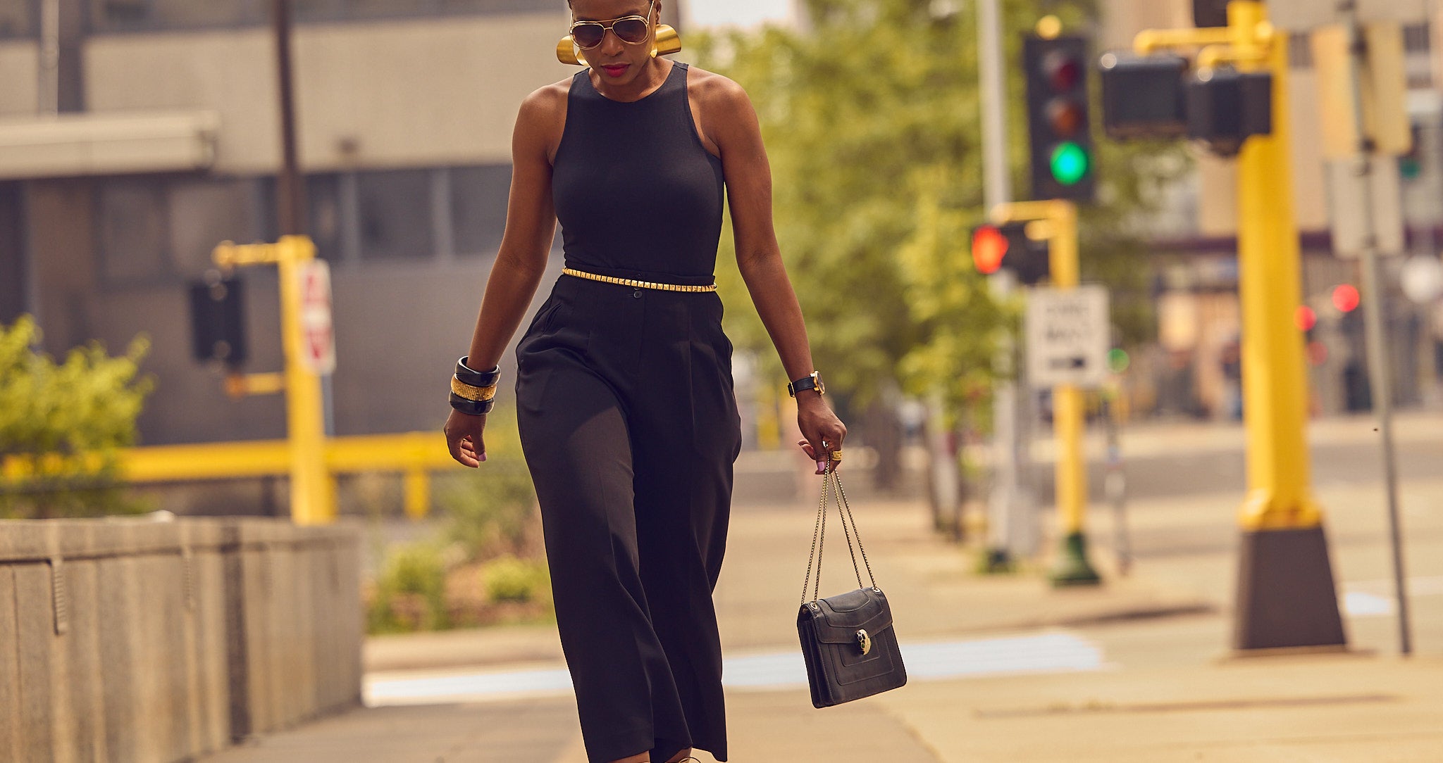 Fashion influencer Farotelle posing while wearing an all-black outfit with gold heeled sandals and gold jewelry.