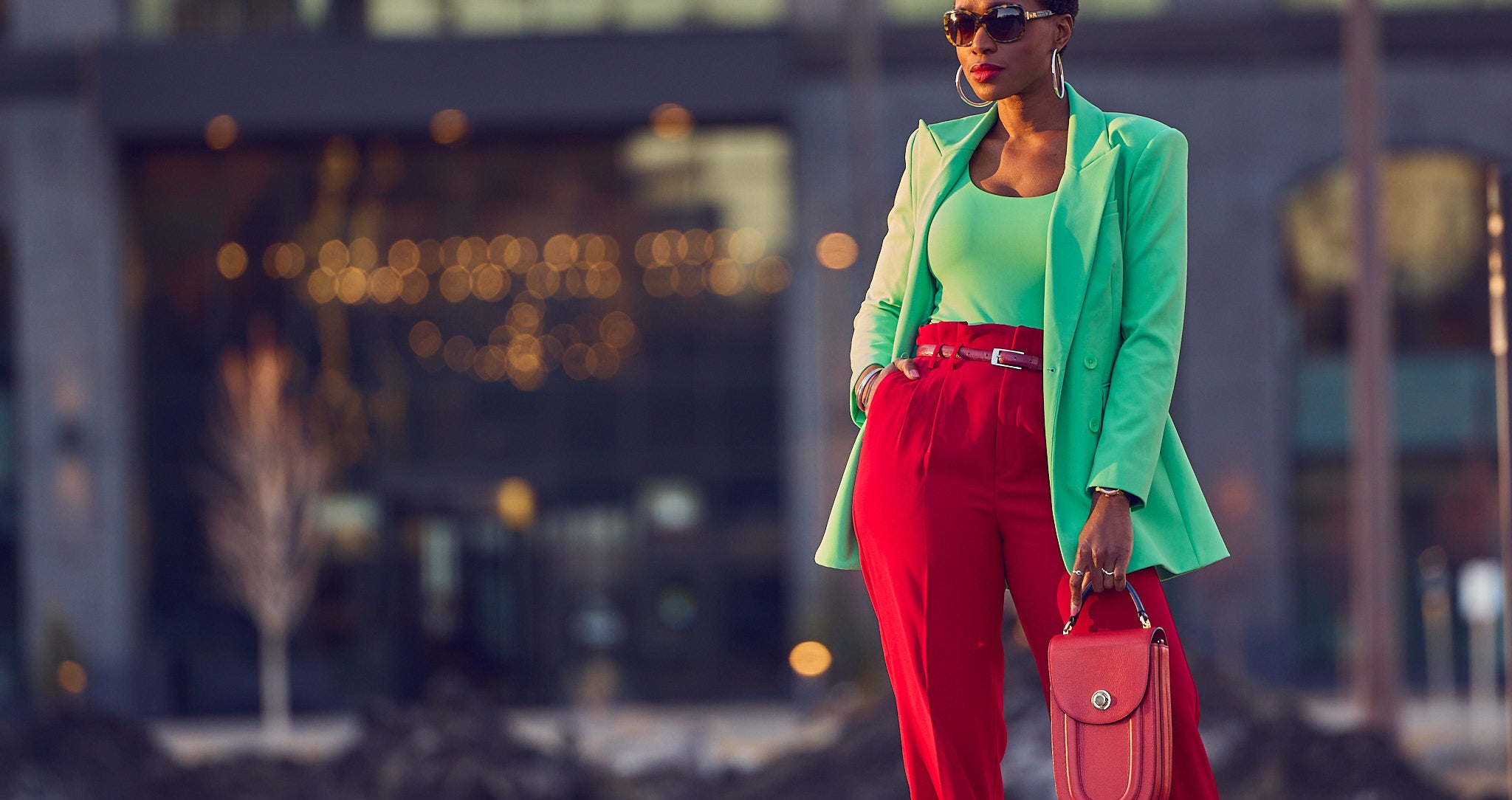 A Fashion editorial photo of style influencer Farotelle wearing a red and green color-blocked outfit. She has on red pants with a fitted green bodysuit and a matching green blazer. She's holding a red handbag and is wearing sunglasses.