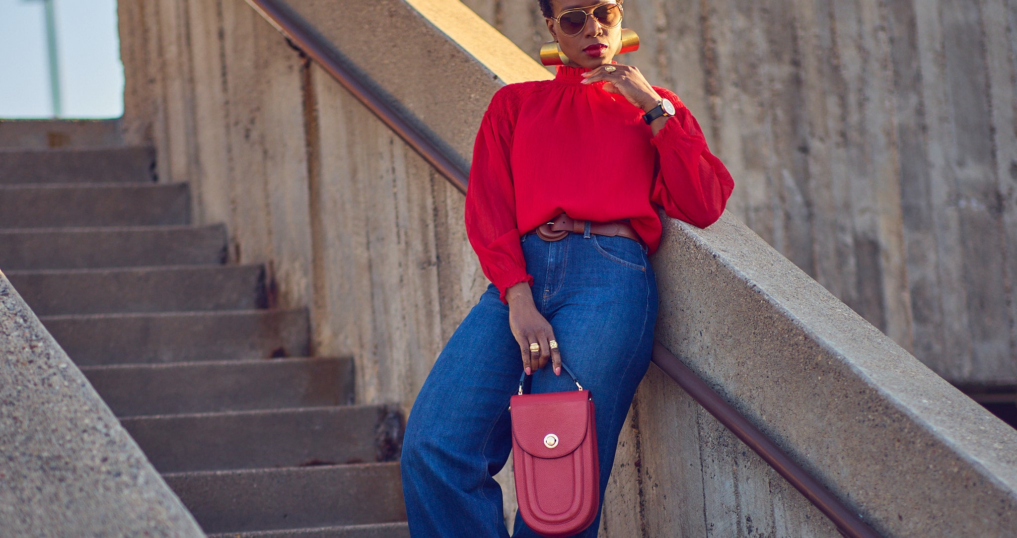 Style influencer Farotelle wearing a red and blue color-blocked outfit. She has on blue wide-leg jeans with a bright red blouse, red pumps, and a red oval-shaped handbag. She's standing next to a wall and is wearing oversized gold earrings.