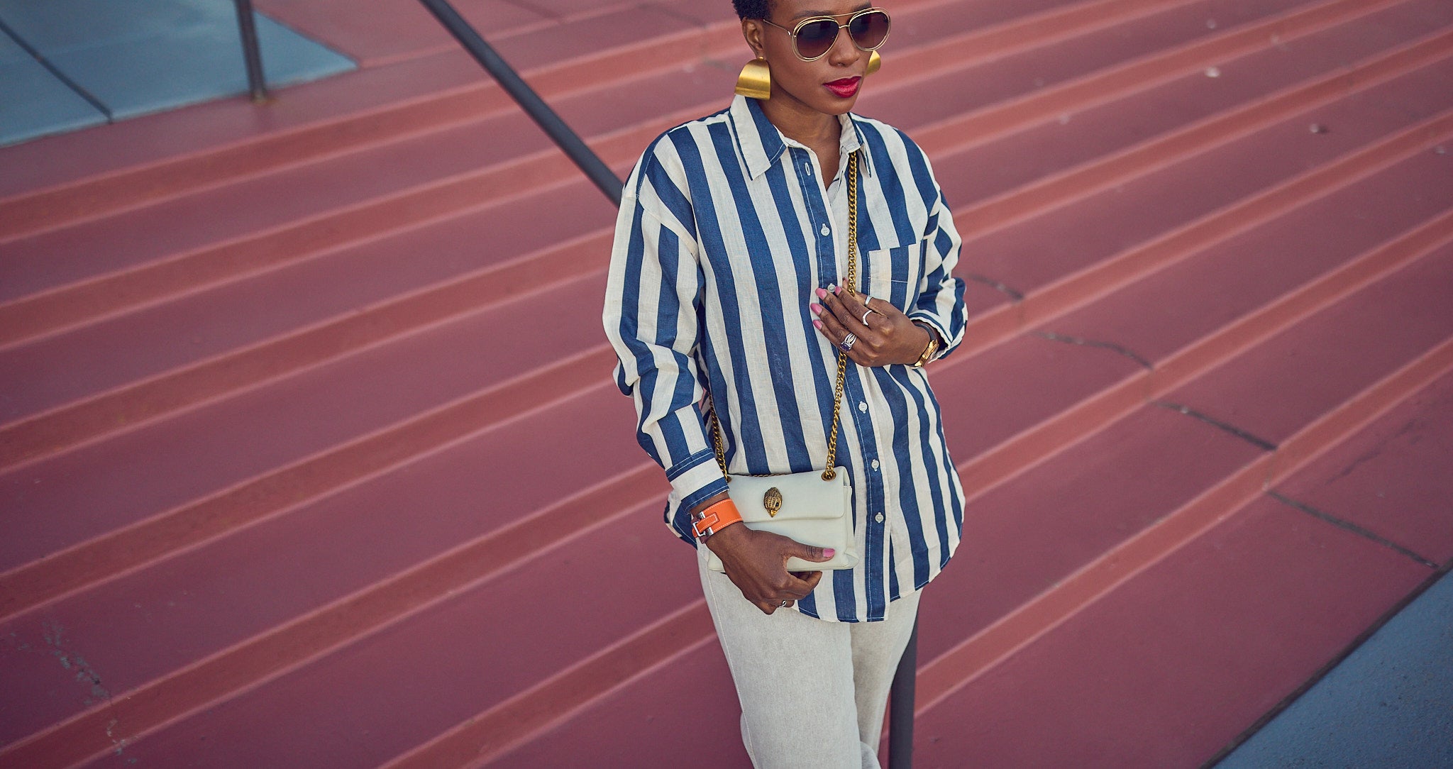 Style influencer Farotelle wearing an oversized blue and beige striped shirt with beige pants. She's wearing sunglasses and a Kurt Geiger chain crossbody handbag. This is an elevated casual Spring look. She is standing against contrastive reddish stairs.